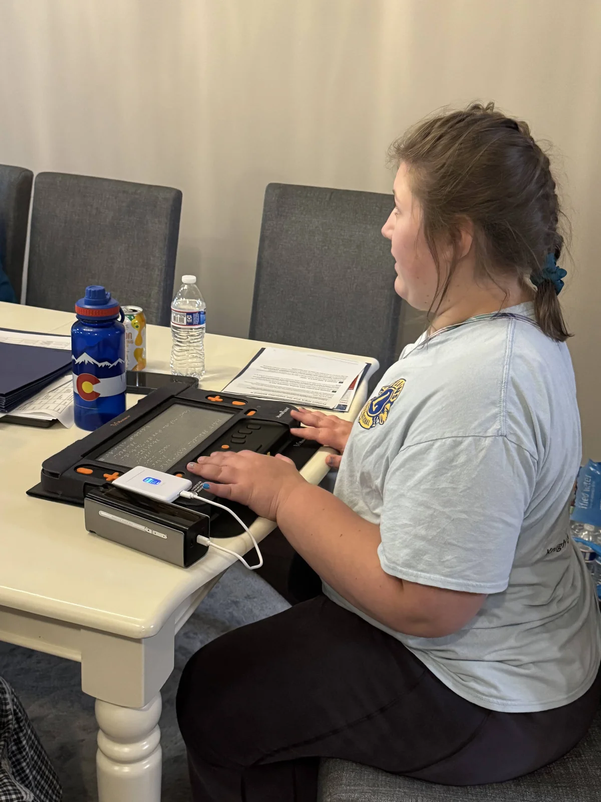Isabelle sits at a table with a laptop and Braille device.