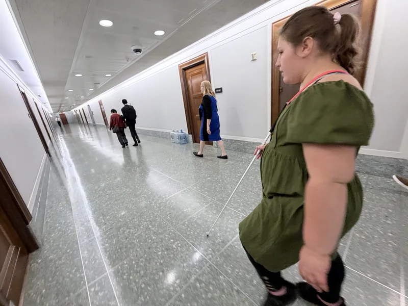 Isabelle, wearing a green dress, travels along a wide corridor with her white cane.