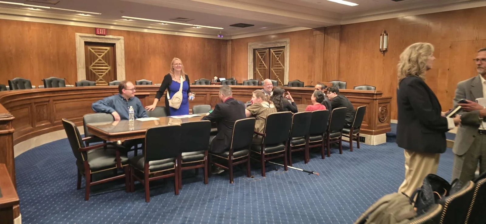 The group gathers around tables in a wood-paneled hearing room.