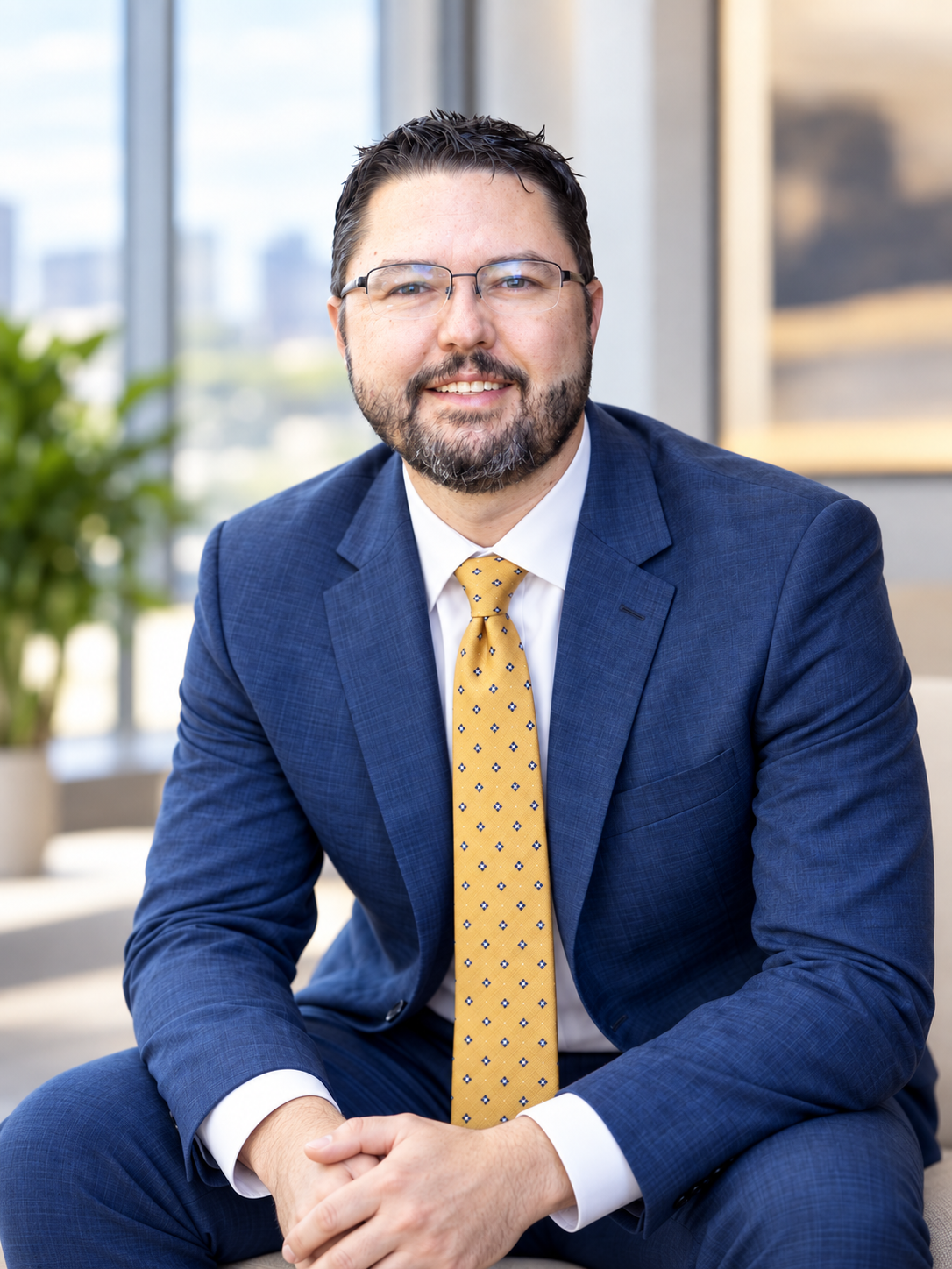 Rob Harris seated indoors, wearing glasses, a blue suit, and a gold patterned tie.