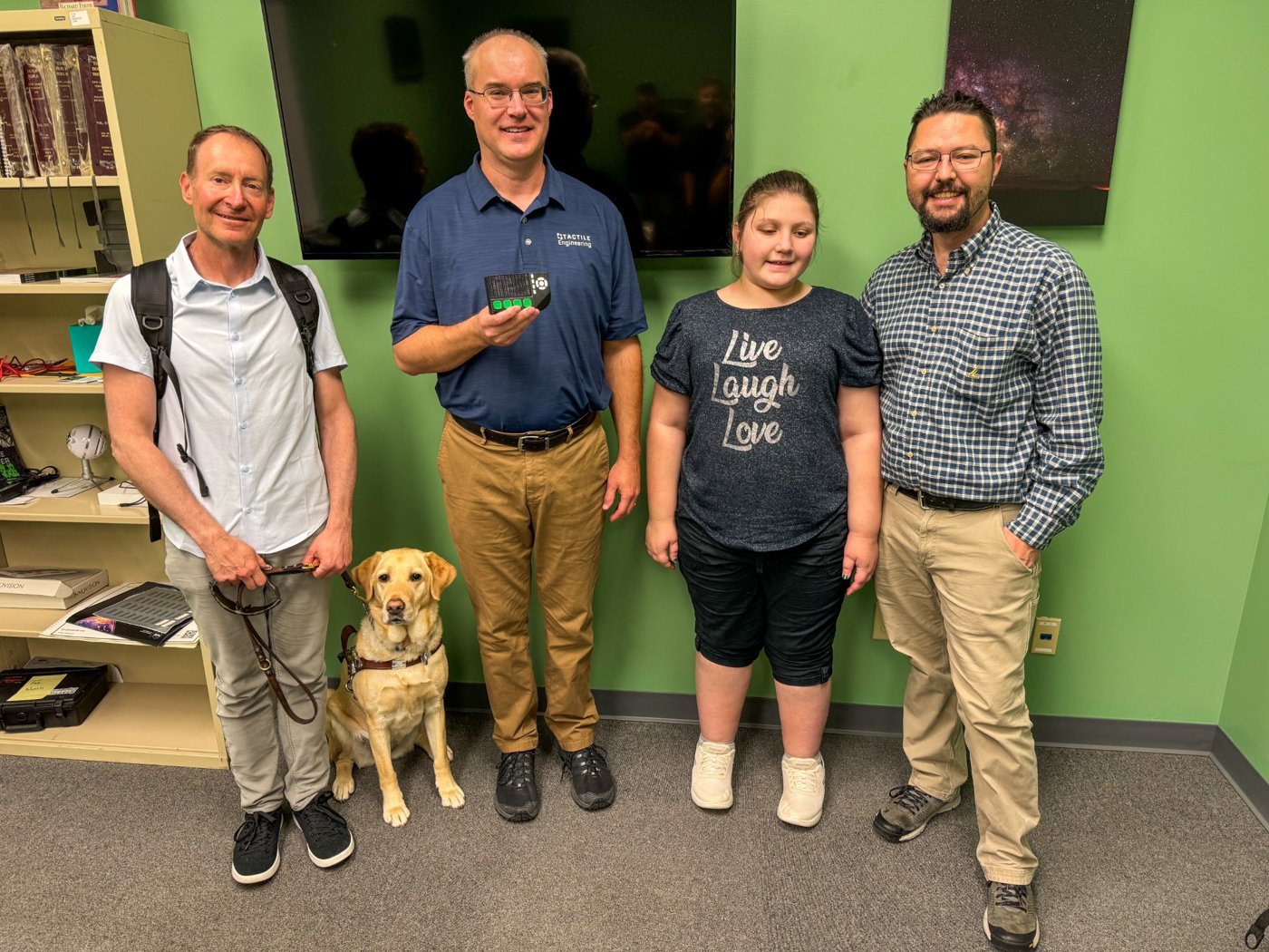 Rob and Isabelle stand with two men and a guide dog; a man wearing a Tactile Engineering shirt holds a tactile device.