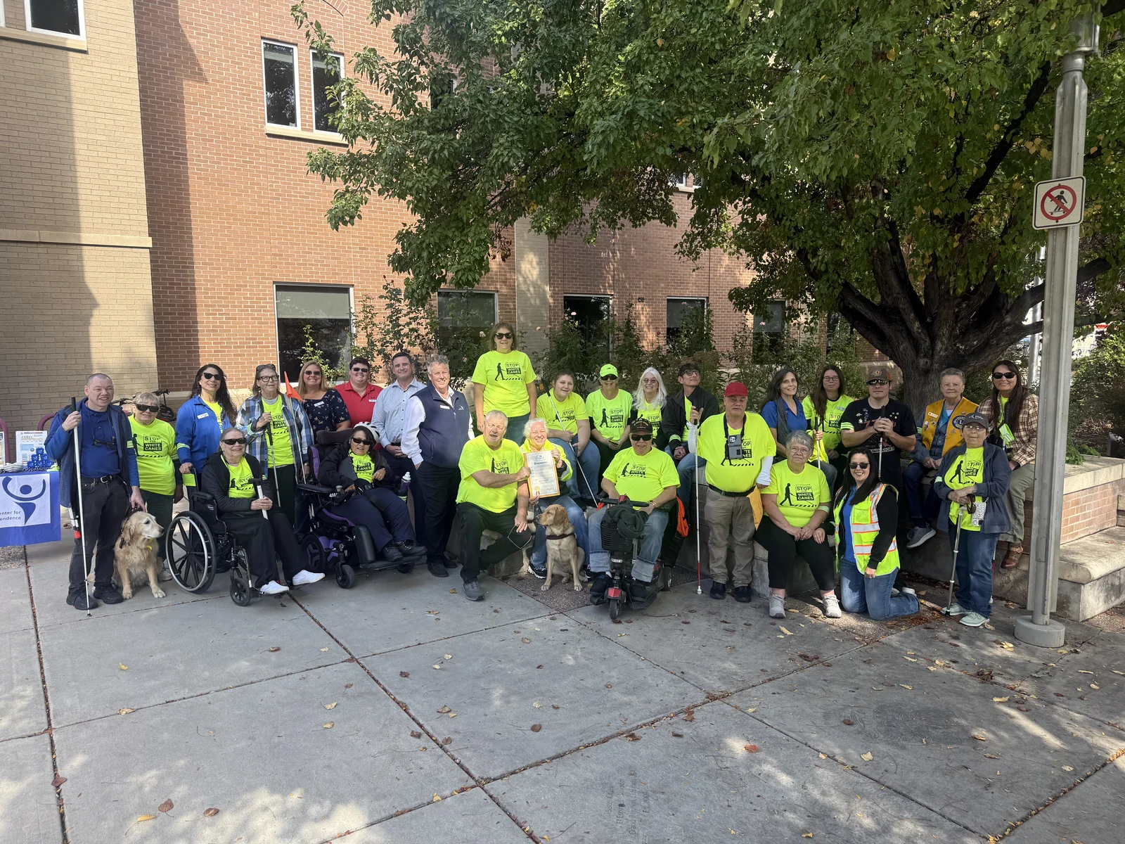 White Cane Day participants gather under a tree in downtown Grand Junction with white canes, guide dogs, wheelchairs, and bright yellow Stop for White Canes shirts.