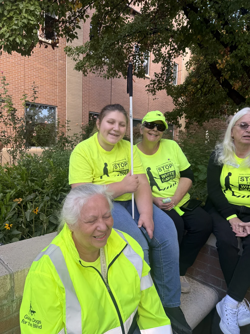 Isabelle sits with fellow participants outdoors; they wear bright yellow shirts and a high-visibility jacket, and Isabelle holds her white cane.