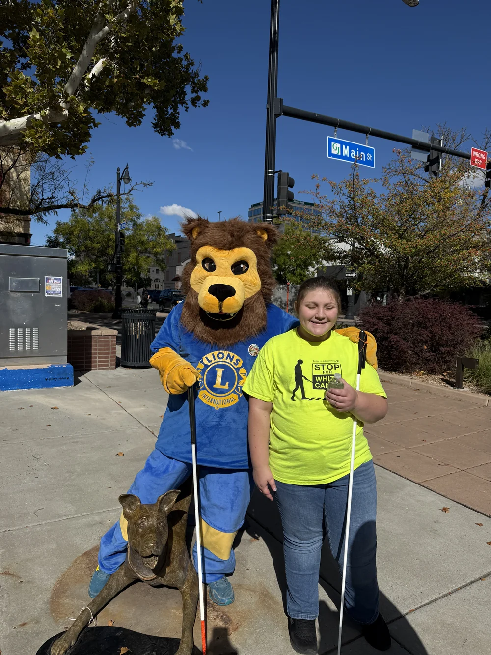 Isabelle smiles beside the Lions Club lion mascot near the Main Street sign, holding her white cane and wearing a Stop for White Canes shirt.